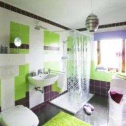 Jasna łazienka po remoncie z zielono-brązowymi akcentami: płytki na ścianach, dywanik, zasłona prysznicowa, umywalka z baterią, toaleta i okno z fioletową zasłoną.